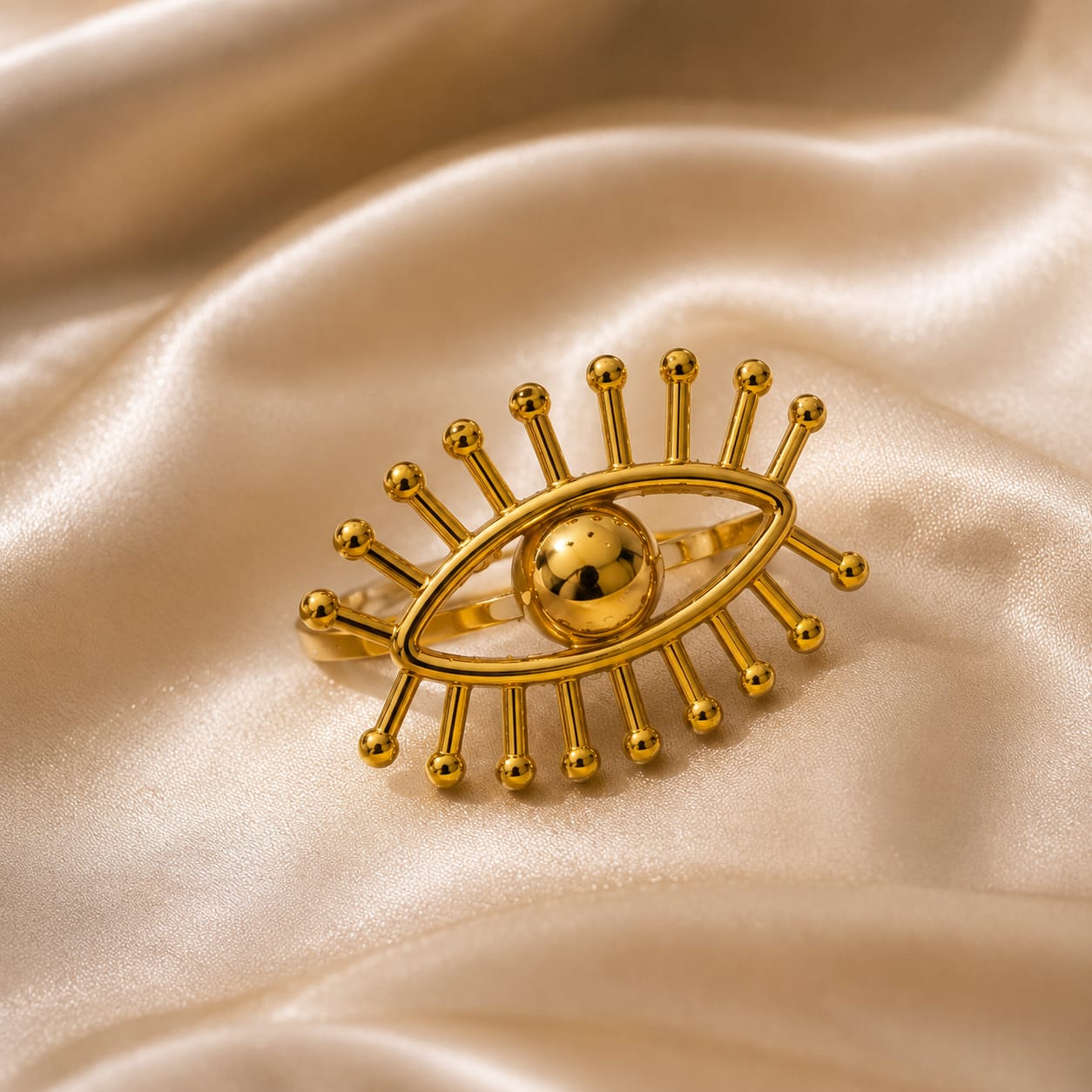 A gold-toned eye-shaped ring with radiating bead-like rays and a polished central sphere, displayed on draped satin fabric.
