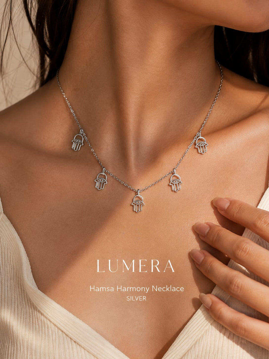 A silver Hamsa hand necklace with five hanging charms, shown on a model's neck and chest. The text 'LUMERA Hanisa Harmony Necklace SILVER' is displayed on the lower chest.