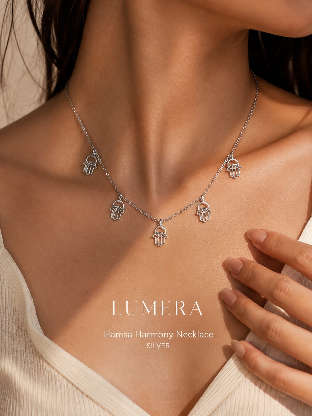 A silver Hamsa hand necklace with five hanging charms, shown on a model's neck and chest. The text 'LUMERA Hanisa Harmony Necklace SILVER' is displayed on the lower chest.