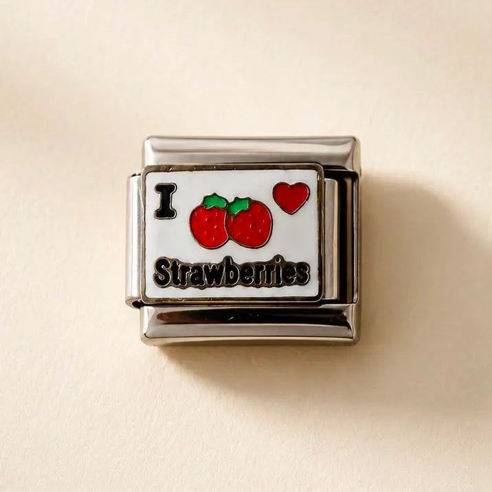 A rectangular Italian charm link for jewelry featuring a white face with red strawberries, a red heart, the letter 'I', and the word 'Strawberries' in black script, set in polished silver-tone metal on a light beige background.