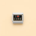 A rectangular silver-toned charm link with a black face, featuring the text 'I' and 'COKE' in gold-tone lettering and a small red heart symbol in the center, shown on a solid pale peach background.