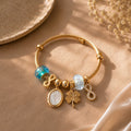 A gold-plated twist bangle bracelet with various charms, including a blue-green bead, a light blue faceted bead, a white oval charm, a gold four-leaf clover charm, and a gold infinity charm, displayed on a beige fabric background.