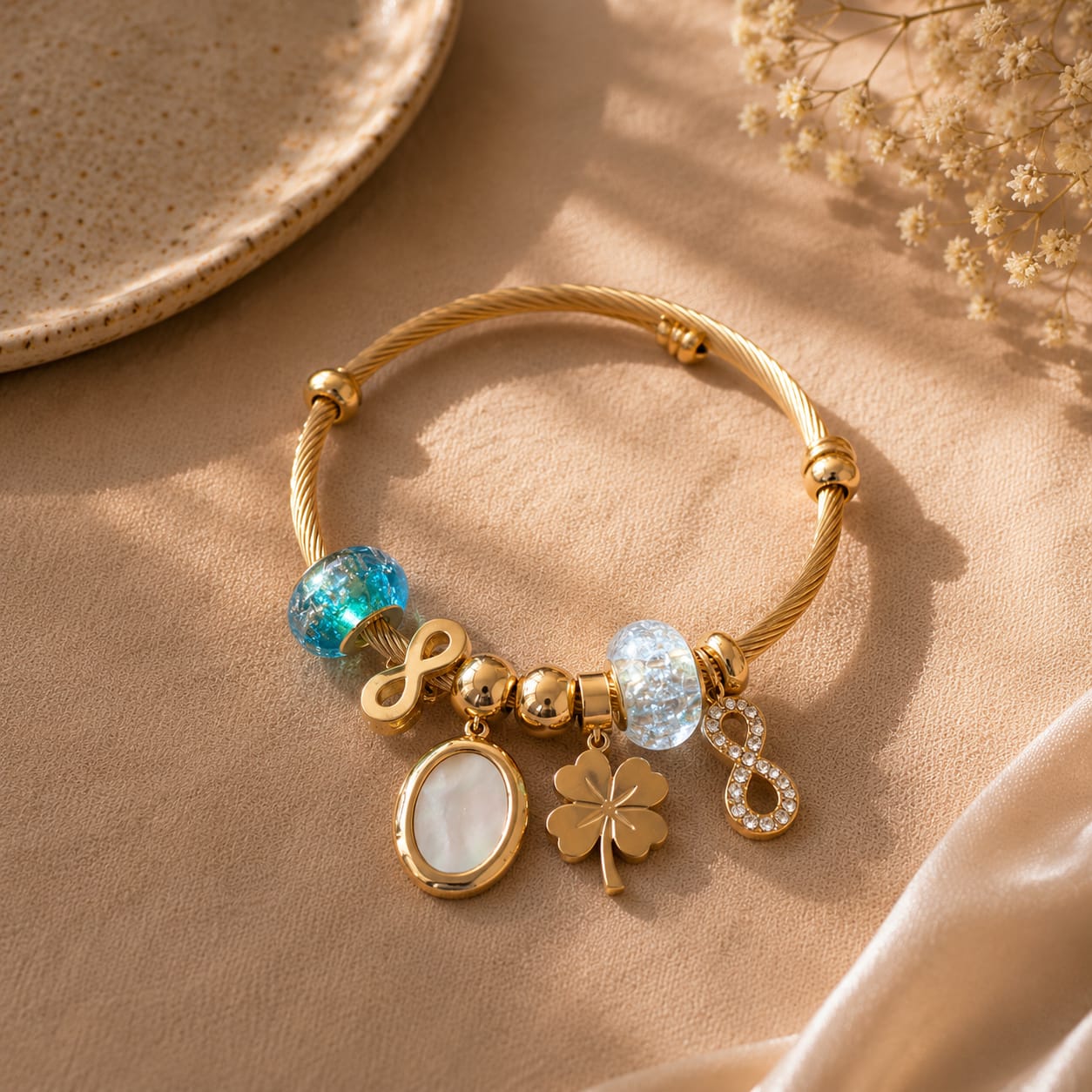 A gold-plated twist bangle bracelet with various charms, including a blue-green bead, a light blue faceted bead, a white oval charm, a gold four-leaf clover charm, and a gold infinity charm, displayed on a beige fabric background.