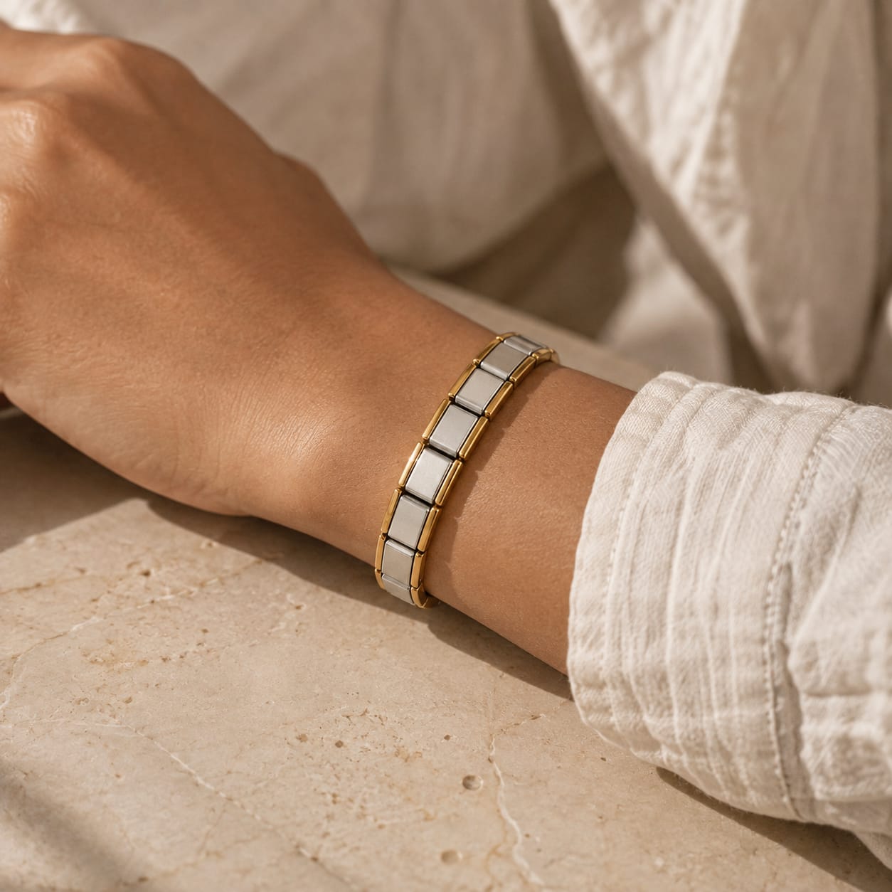 A gold and white ceramic link bracelet worn on a person's wrist, shown in a warm, sunlit lifestyle setting with a white textured shirt.