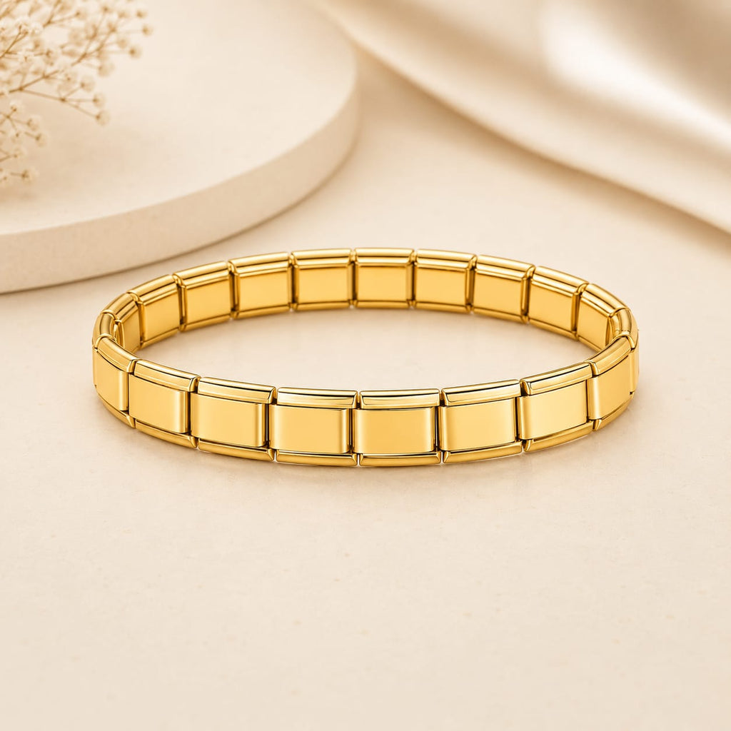 A gold-toned stretch bracelet made of rectangular link segments, photographed on a warm cream surface with soft decorative props in the background.
