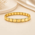 A gold-toned stretch bracelet made of rectangular link segments, photographed on a warm cream surface with soft decorative props in the background.
