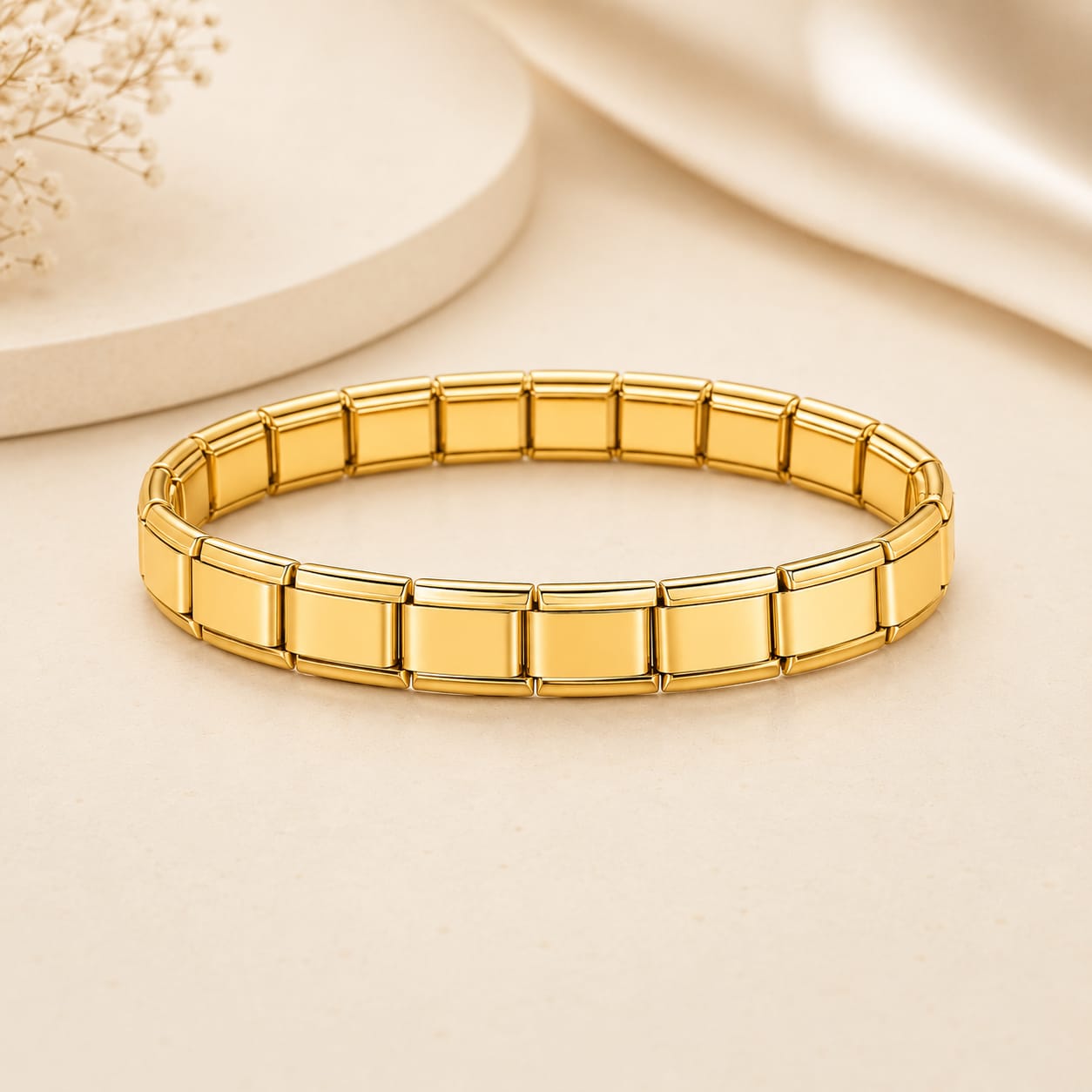 A gold-toned stretch bracelet made of rectangular link segments, photographed on a warm cream surface with soft decorative props in the background.