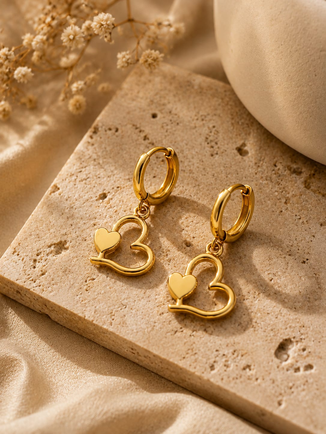 A pair of gold hoop earrings with open heart-shaped dangling pendants, styled on a textured beige stone slab with dried flowers and soft neutral fabric in the background.