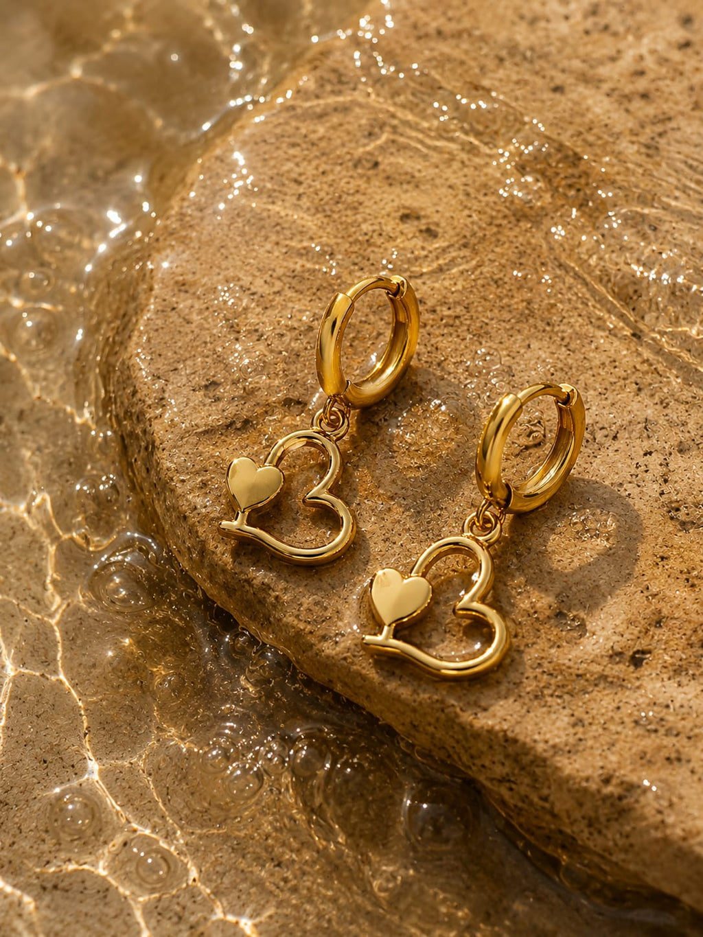 A pair of gold hoop earrings with dangling open heart charms, resting on a wet brown stone with water droplets around them.