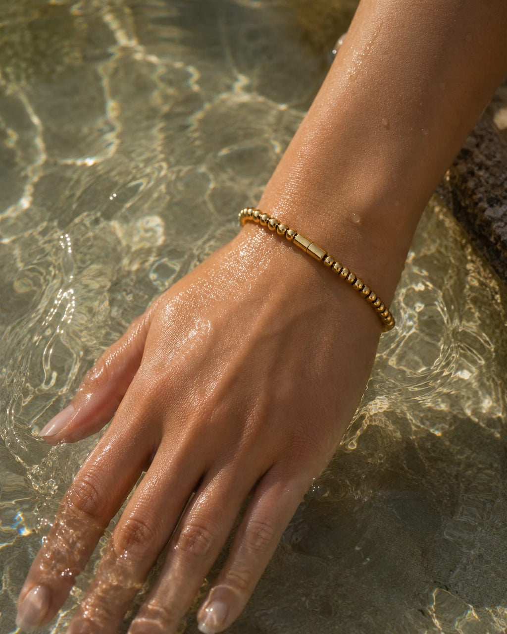 A gold beaded bracelet with a central polished bar, worn on a person's wrist as their hand rests in shallow water.