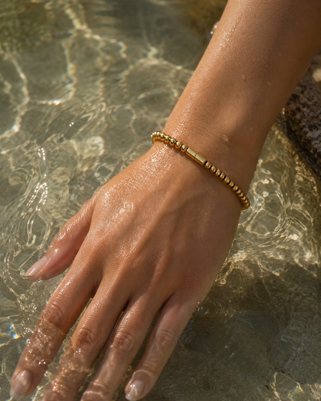 A gold beaded bracelet with a central polished bar, worn on a person's wrist as their hand rests in shallow water.