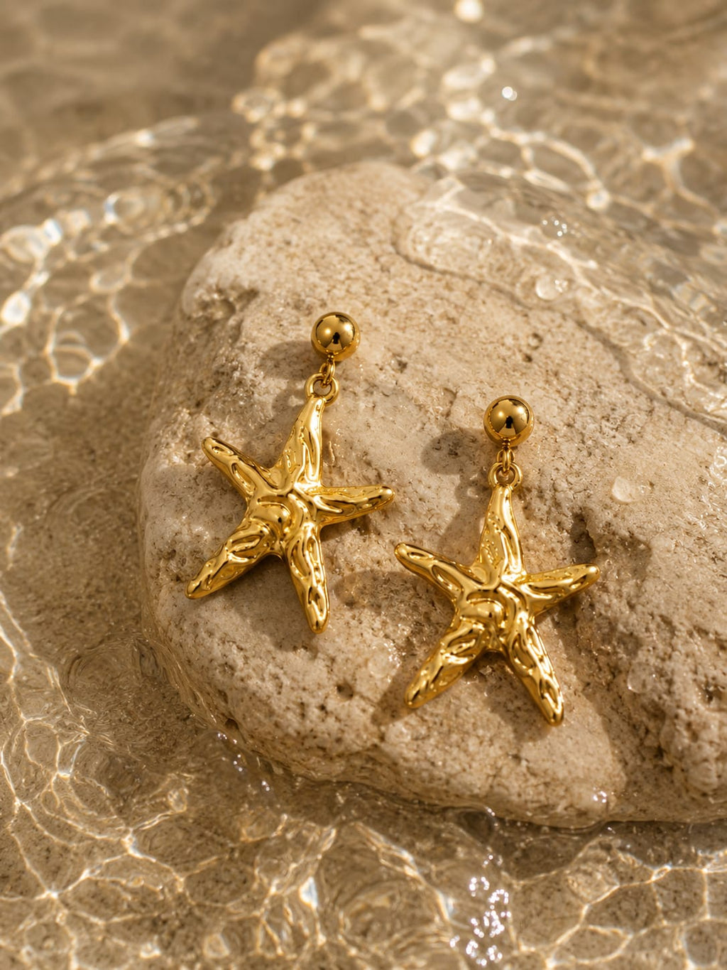 A pair of gold starfish-shaped drop earrings resting on a smooth stone, with shallow water and sunlight ripples in the background.