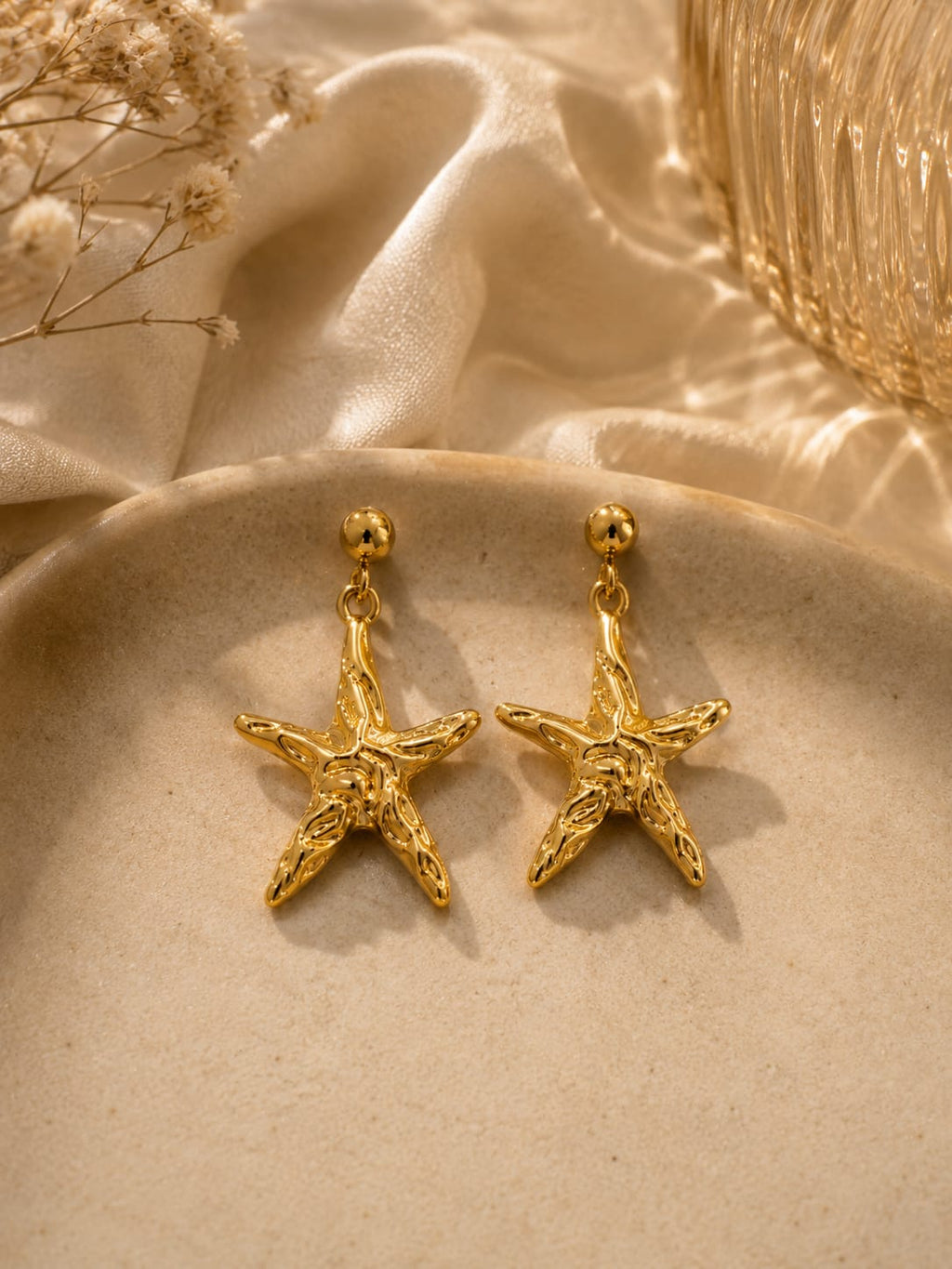 A pair of gold starfish-shaped dangle earrings displayed on a beige ceramic dish with draped satin fabric and dried flowers in the background.