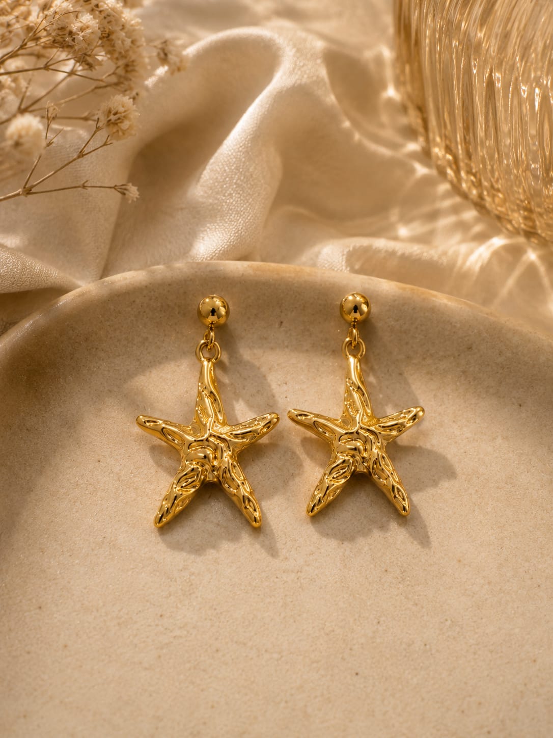 A pair of gold starfish-shaped dangle earrings displayed on a beige ceramic dish with draped satin fabric and dried flowers in the background.