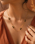 A close-up lifestyle shot of a model wearing a delicate gold chain necklace adorned with three small gold bee charms, each holding a black heart-shaped stone. The model is wearing a rust-colored top, and the background is a warm, softly lit brown.