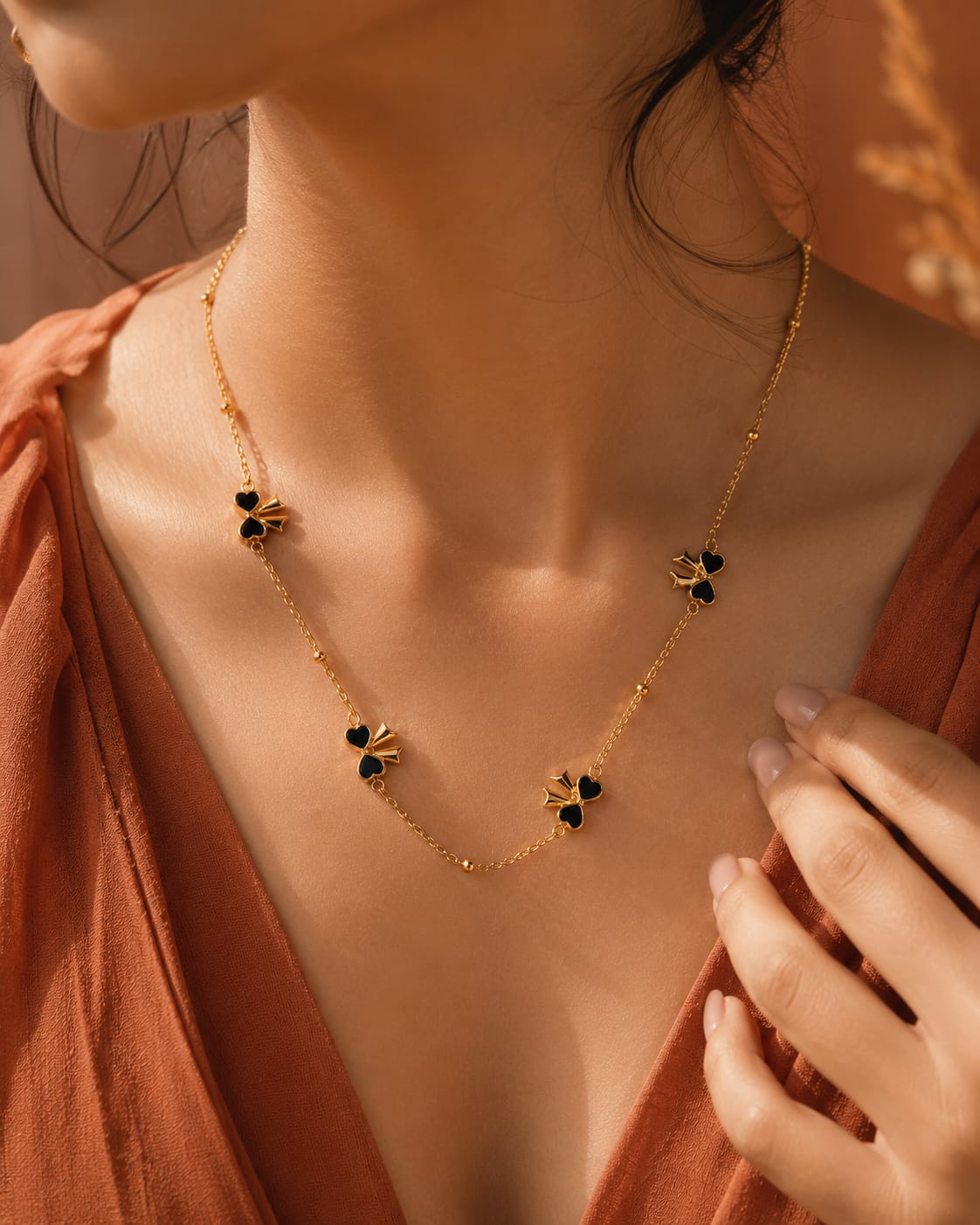A close-up lifestyle shot of a model wearing a delicate gold chain necklace adorned with three small gold bee charms, each holding a black heart-shaped stone. The model is wearing a rust-colored top, and the background is a warm, softly lit brown.