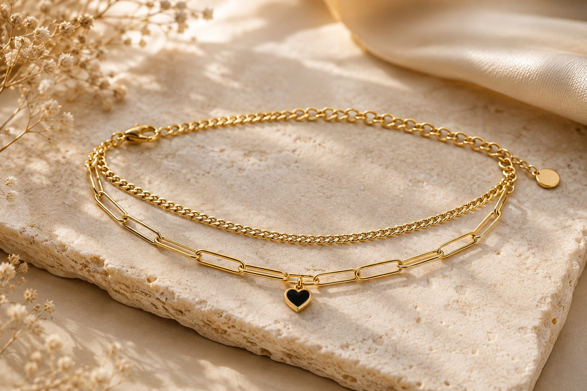 A gold double-layer necklace with a fine chain and a paperclip chain featuring a small heart-shaped charm with a black center, displayed on a textured stone slab with dried flowers.