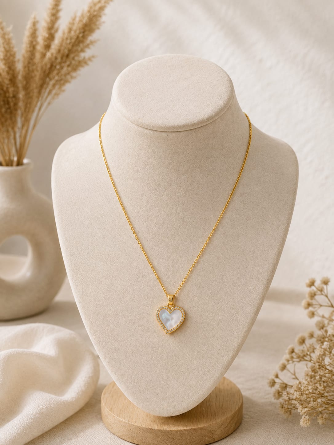 A delicate gold chain necklace with a heart-shaped pendant featuring a white mother-of-pearl center and a gold border, displayed on a cream-colored jewelry bust with dried floral accents.