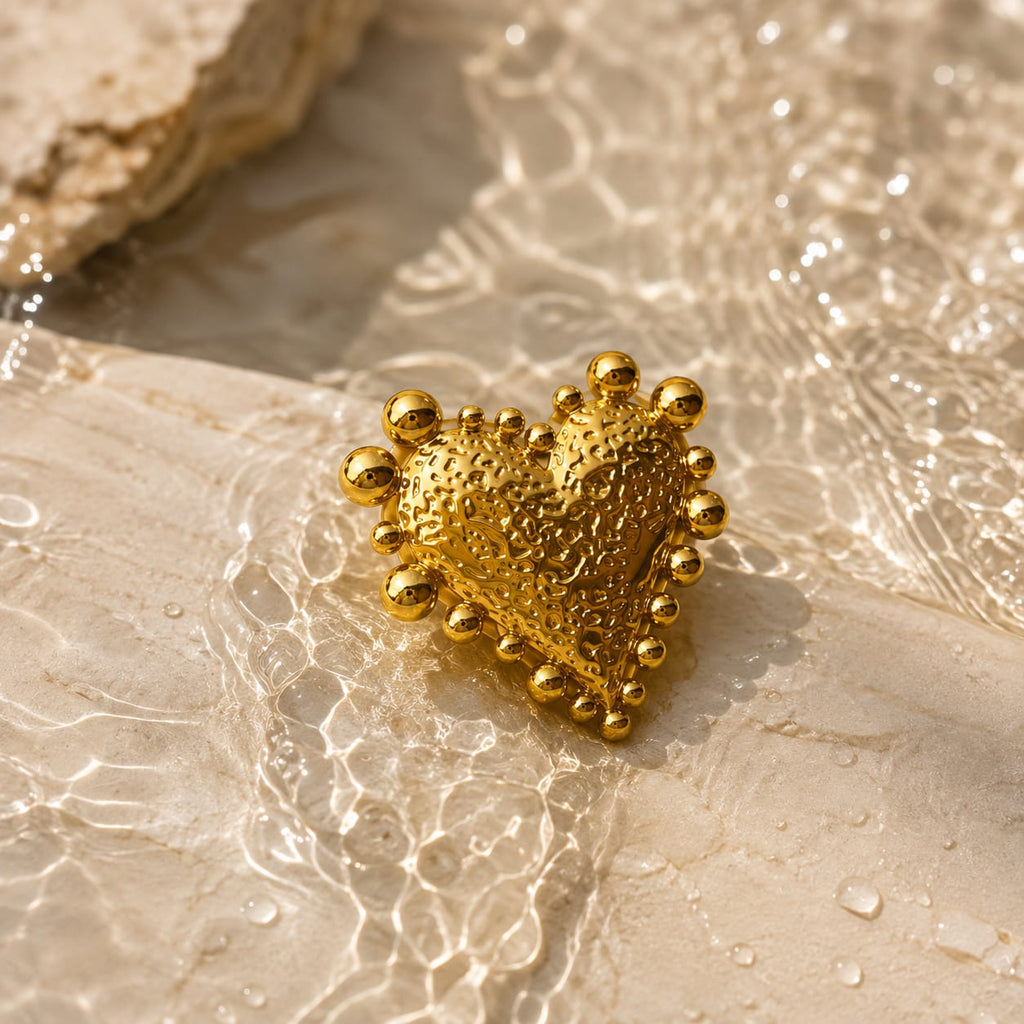 A textured gold heart-shaped pendant with raised beaded edges, resting on a warm beige stone surface with water droplets and light reflections.