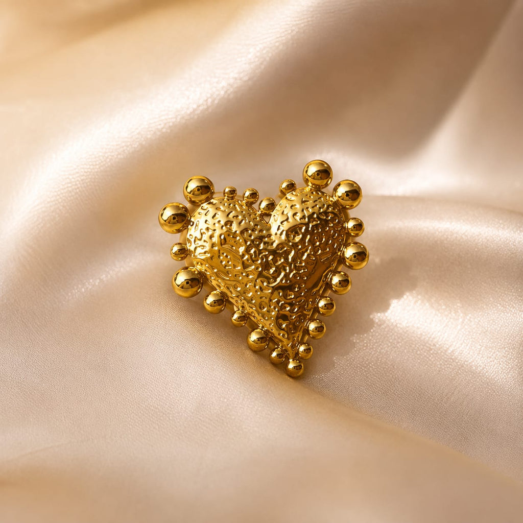 A textured gold heart-shaped brooch with raised beaded edges, displayed on a draped cream satin fabric background.