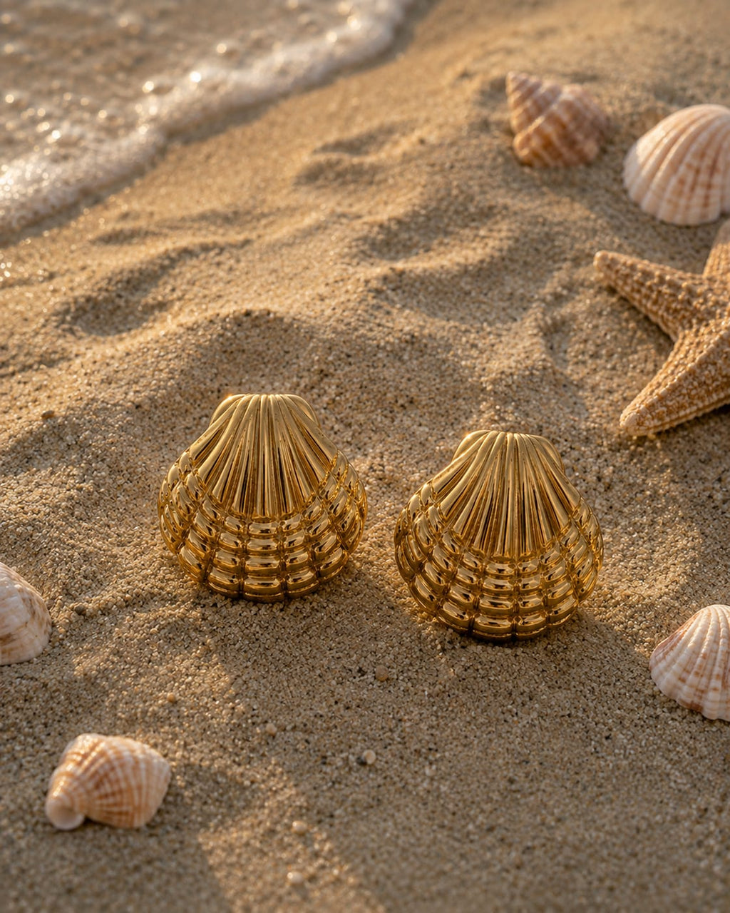 Two gold-toned seashell-shaped jewelry pieces displayed on sandy beach terrain alongside real starfish and natural seashells under warm sunlight.