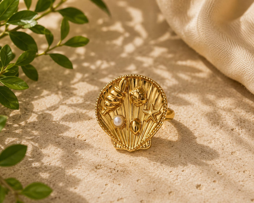 A gold seashell-shaped ring with starfish, shell, and pearl details, styled on a sandy surface with dappled sunlight, green leaves, and a cream fabric background.