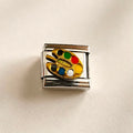 A rectangular silver-tone charm link featuring an embossed gold paint palette with colorful paint dots in green, red, yellow, blue, black, and white, shown on a textured off-white background.