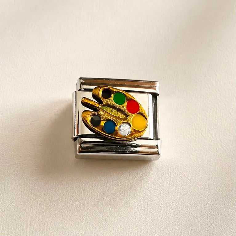 A rectangular silver-tone charm link featuring an embossed gold paint palette with colorful paint dots in green, red, yellow, blue, black, and white, shown on a textured off-white background.