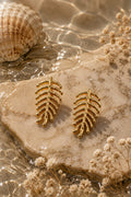 A pair of gold leaf-shaped stud earrings displayed on a beige stone slab with water, a seashell, and dried flowers around them.