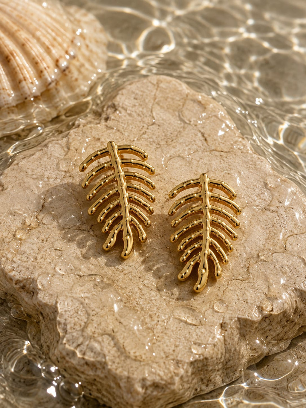 A pair of gold-toned leaf-shaped stud earrings displayed on a textured stone slab with water and a seashell in the background.