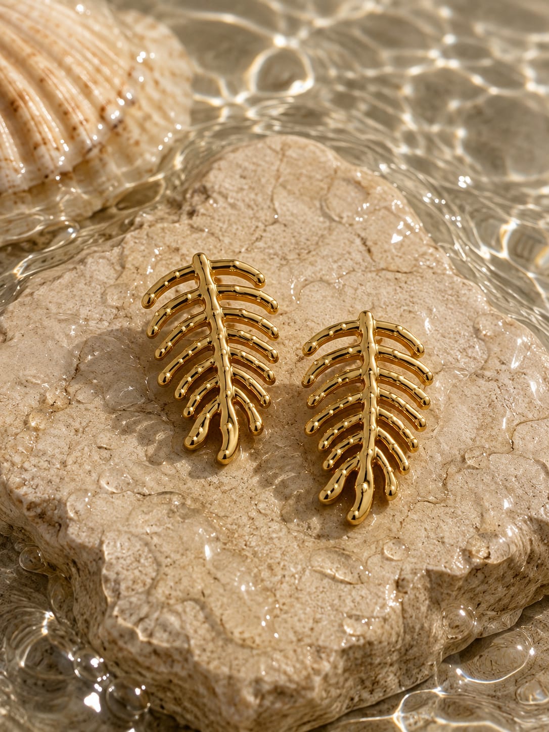 A pair of gold-toned leaf-shaped stud earrings displayed on a textured stone slab with water and a seashell in the background.