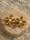 A pair of gold flower-shaped stud earrings with white pearl centers, resting on a beige stone over rippling water.