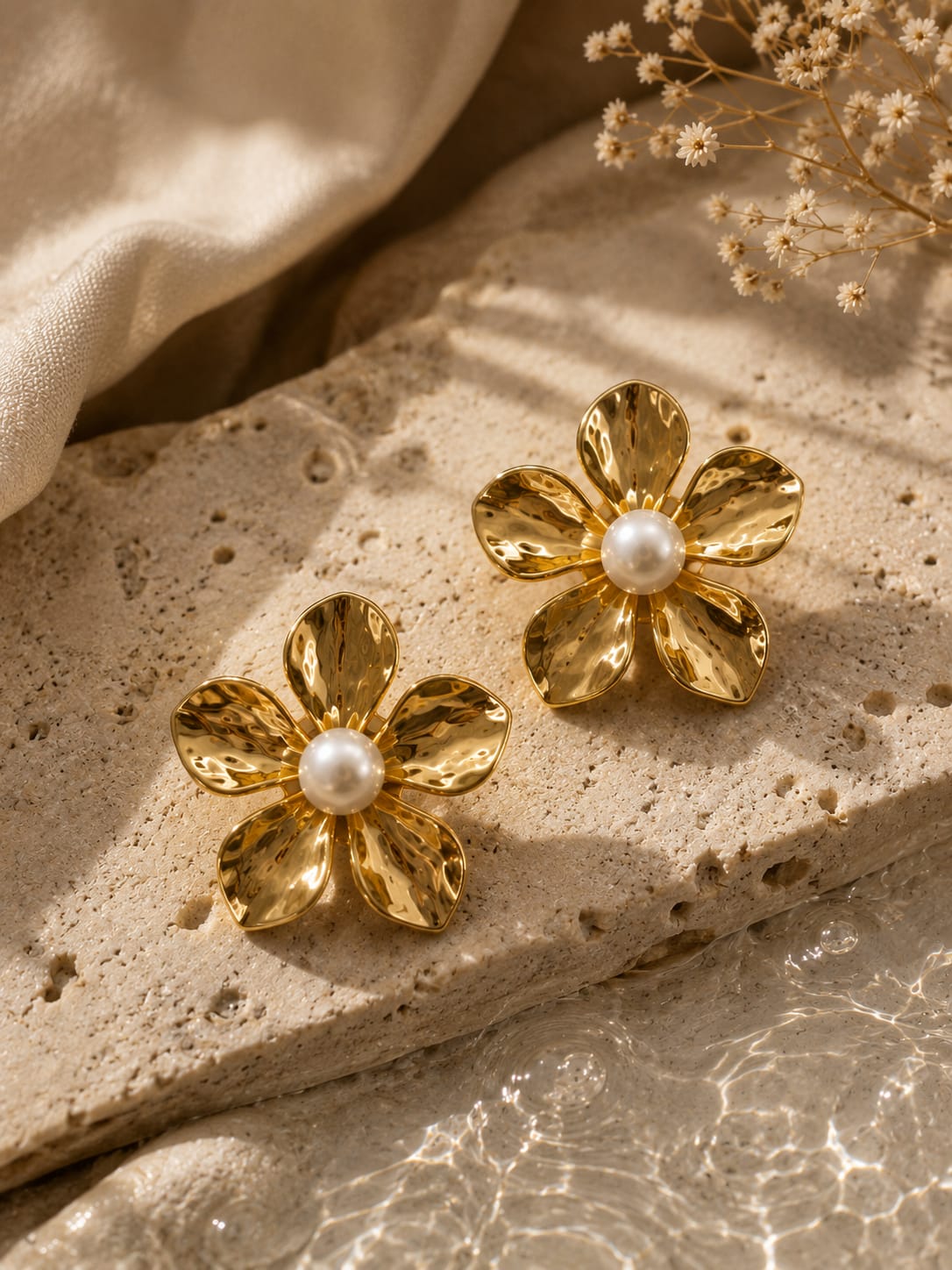 A pair of gold flower-shaped stud earrings with white pearl centers, displayed on a textured beige stone slab with draped satin fabric and dried flowers in the background.