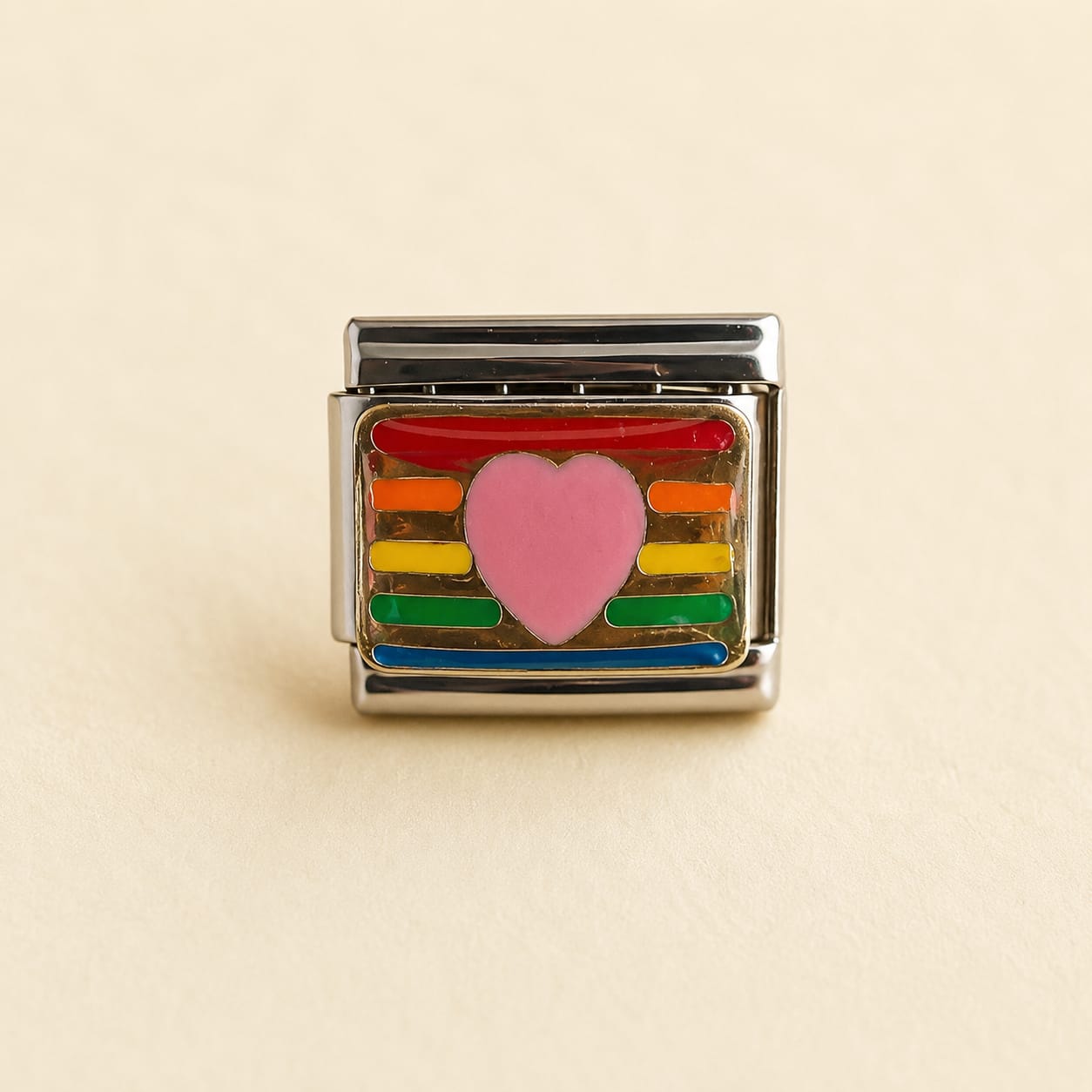 A rectangular silver-toned charm link featuring a pink heart in the center, surrounded by horizontal rainbow stripes in red, orange, yellow, green, and blue, set against a light beige background.