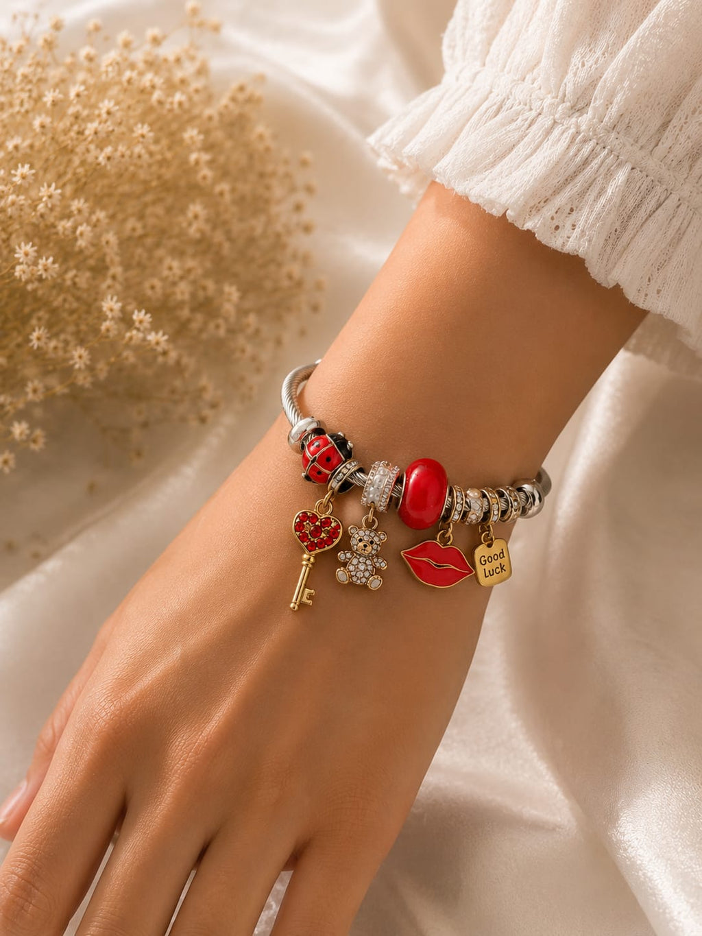 A close-up of a person's wrist wearing a silver charm bracelet with red, gold, and white details, styled with a white sleeve and dried flowers on a white satin background.