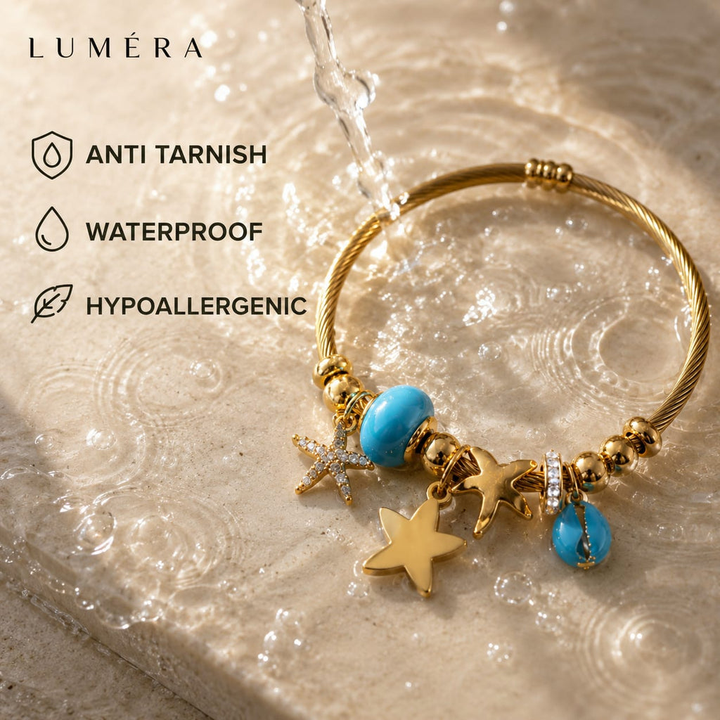Gold-tone bangle bracelet with turquoise blue beads, starfish charms, and a sea star charm, shown under running water on a textured stone surface. Text on the left reads LUMÉRA and lists anti tarnish, waterproof, and hypoallergenic features.