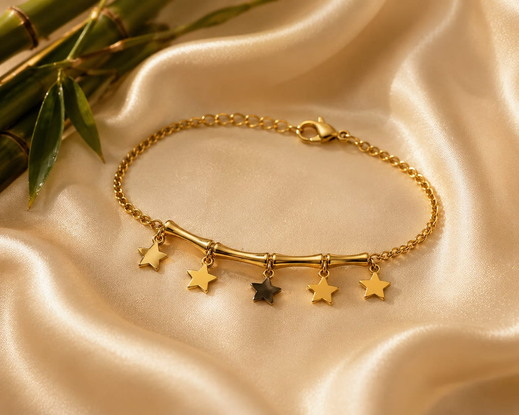 A gold chain bracelet with a curved bar and five dangling star charms, including one dark star, displayed on draped champagne satin fabric with bamboo leaves in the corner.