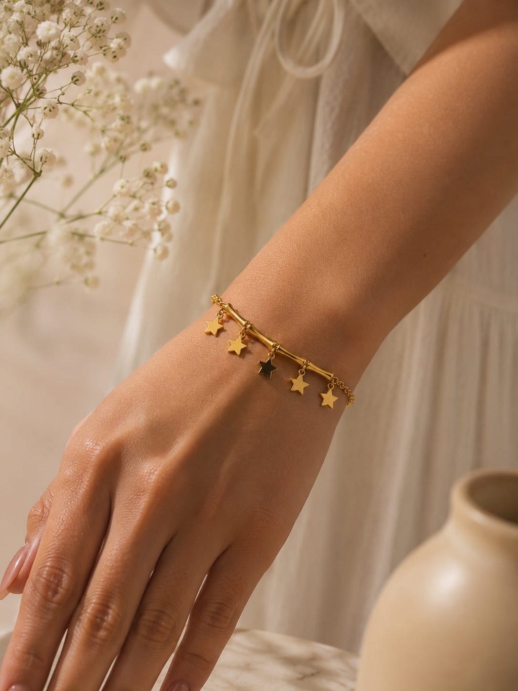A delicate gold bracelet with dangling star charms worn on a woman's wrist, styled with a soft white dress and baby's breath flowers in the background.