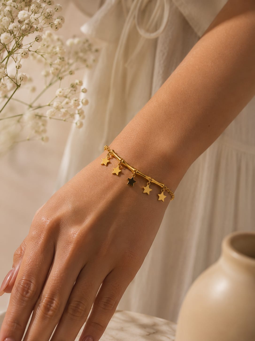 A delicate gold bracelet with dangling star charms worn on a woman's wrist, styled with a soft white dress and baby's breath flowers in the background.