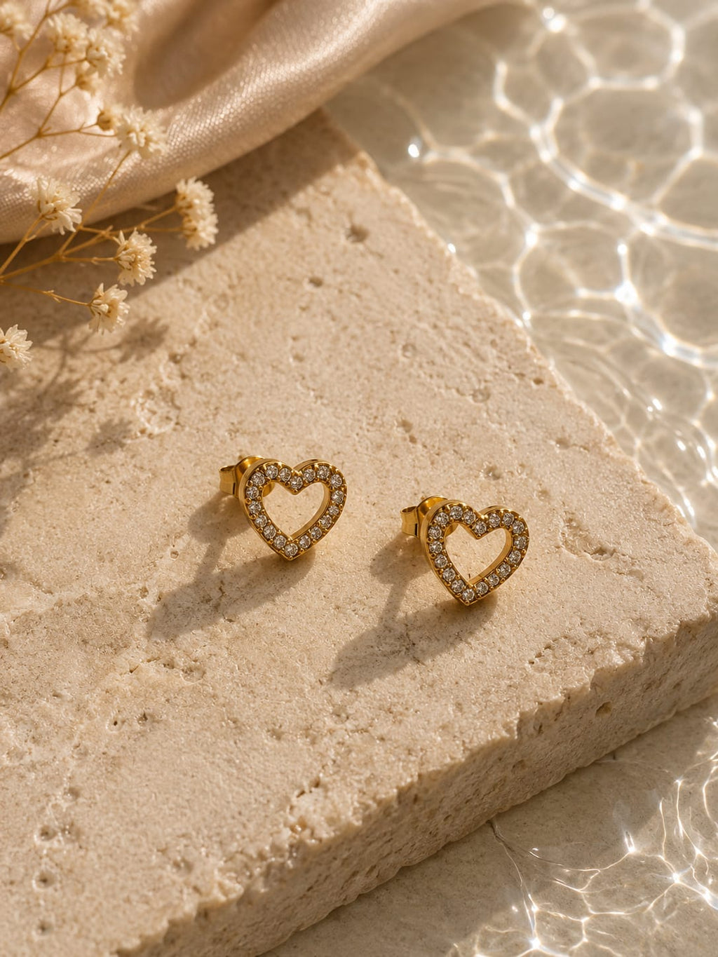 A pair of gold heart-shaped stud earrings with clear crystal accents, displayed on a textured beige stone block with dried flowers and soft sunlight.