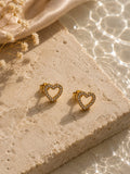 A pair of gold heart-shaped stud earrings with clear crystal accents, displayed on a textured beige stone block with dried flowers and soft sunlight.