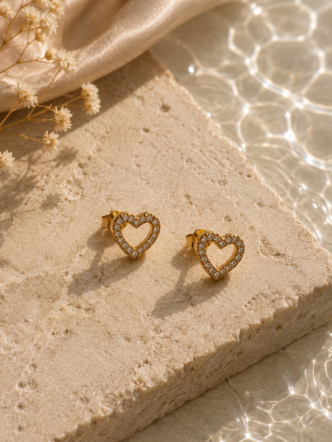 A pair of gold heart-shaped stud earrings with clear crystal accents, displayed on a textured beige stone block with dried flowers and soft sunlight.