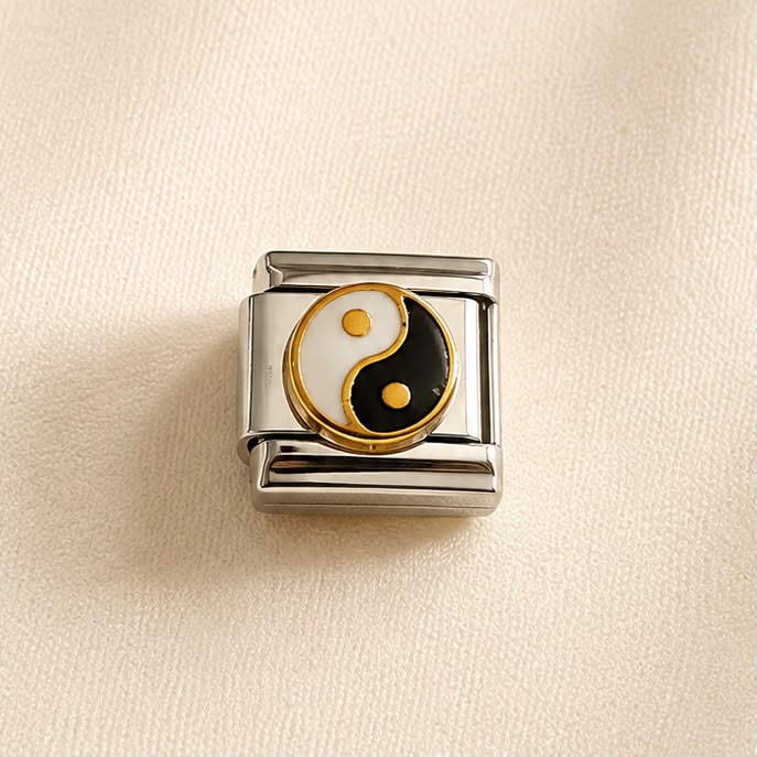 A square silver-toned metal charm link featuring a central Yin Yang symbol with white, black, and gold enamel details, set against a textured beige background.