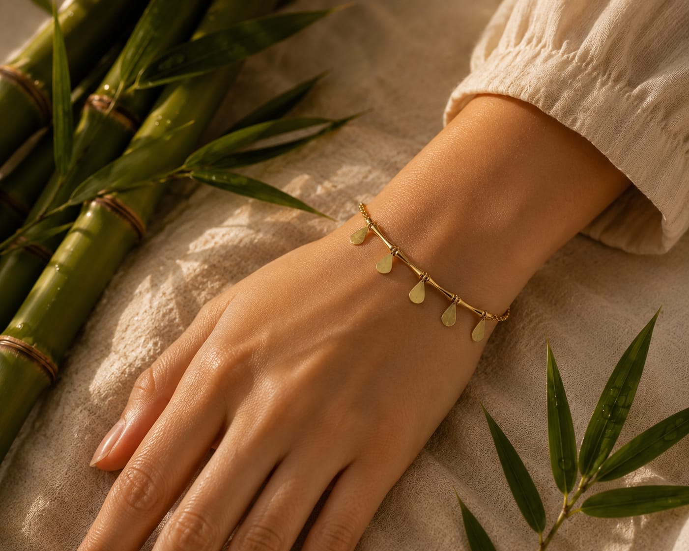 A delicate gold chain bracelet with small teardrop-shaped charms worn on a person's wrist, shown in a styled flat-lay with bamboo leaves and a beige fabric background.