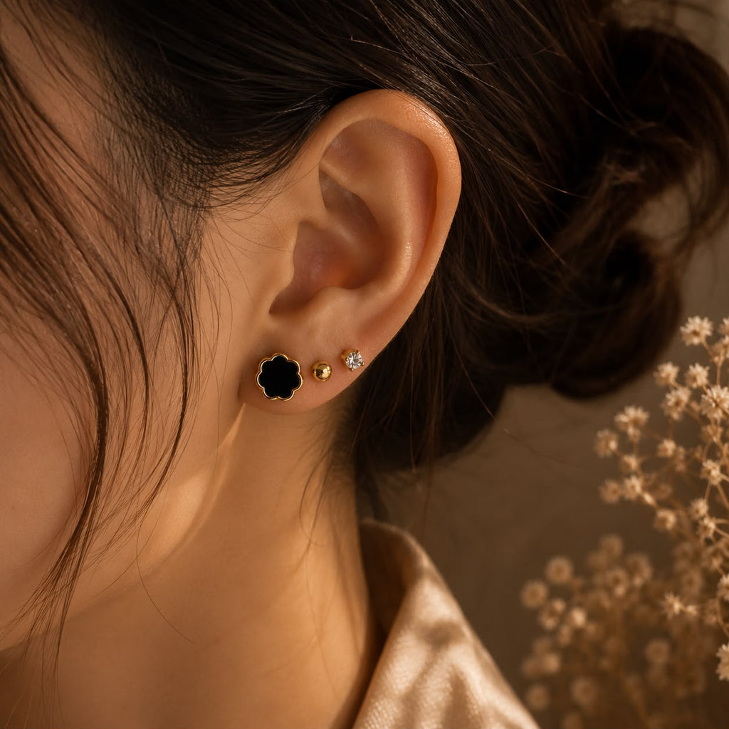 Close-up of a person's ear wearing three earrings: a black flower-shaped stud with a gold border, a small gold bead stud, and a clear crystal stud. The background is warm brown with dried flowers.